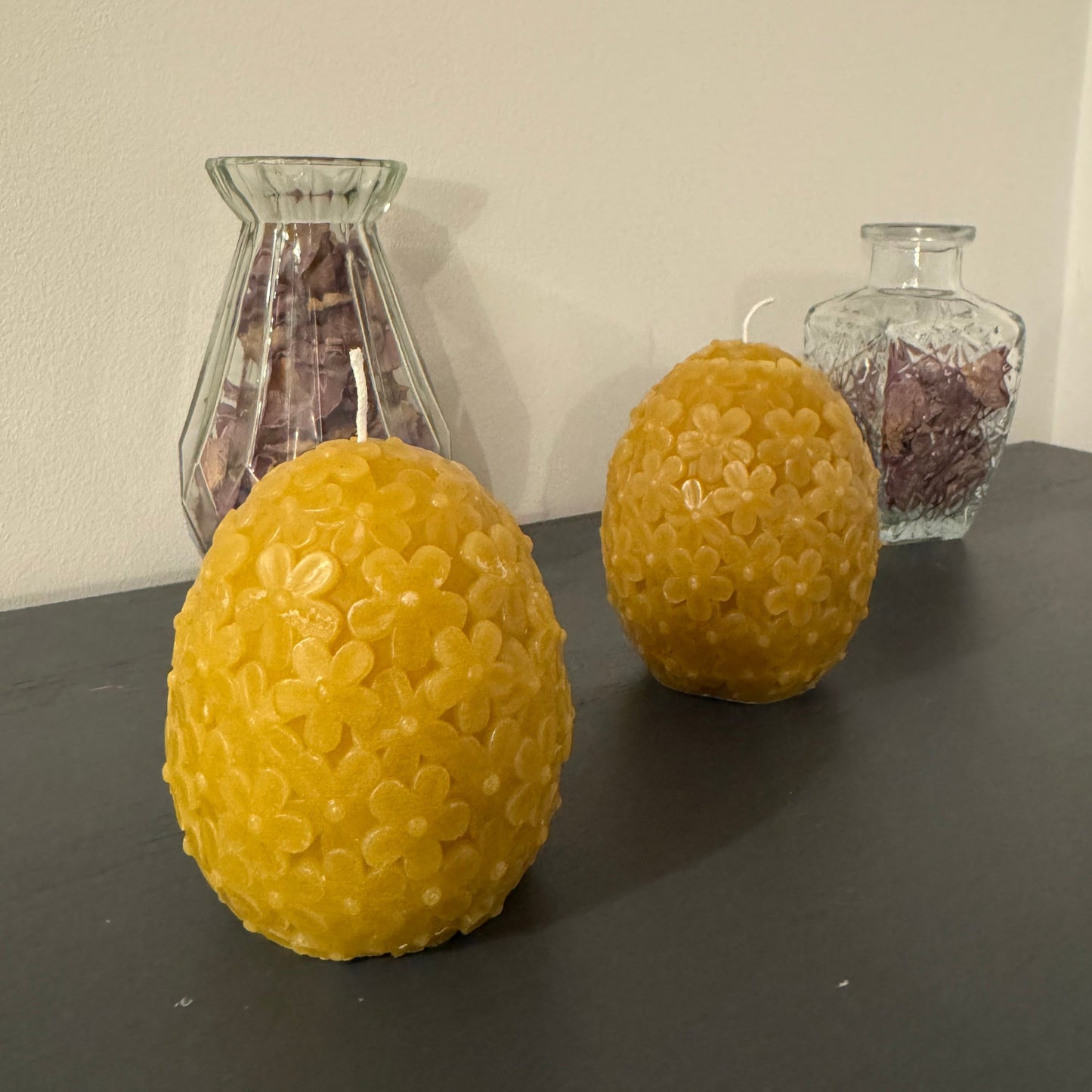 2 floral candles in an egg shape sit on a grey sideboard, with some dried petals behind . It creates a homely, cosy environment
