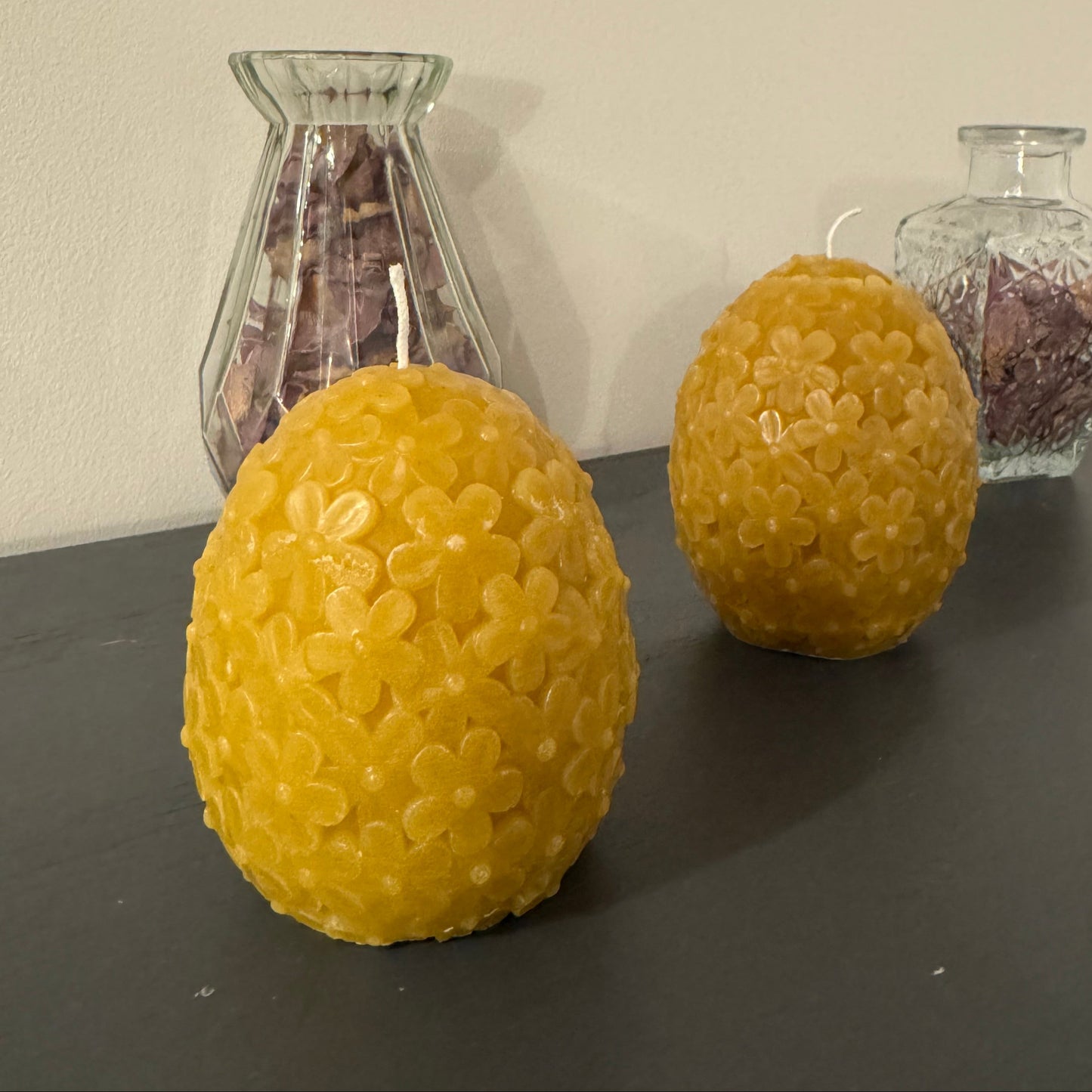 2 egg shaped, dome shaped beeswax candle, with floral detailing in front of 2 vases with dried petals