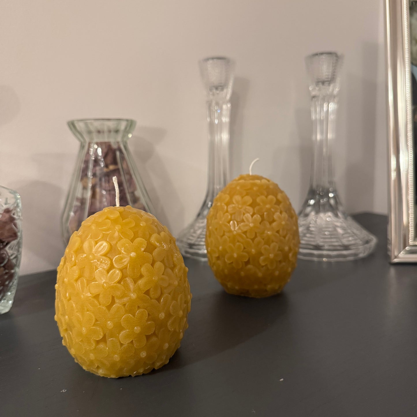 2 floral candles in an egg shape sit on a grey sideboard, with some dried petals behind and picturesque glass candlesticks. It creates a homely, cosy environment