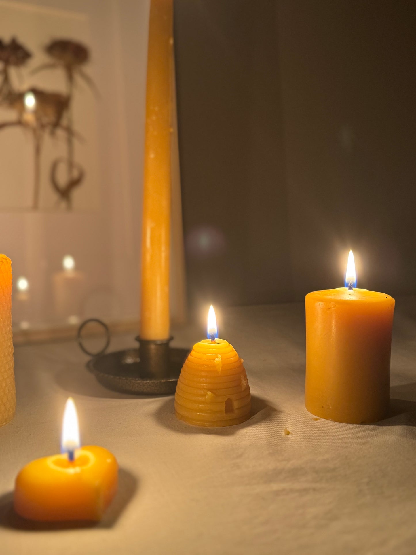 Four handcrafted beeswax candles displayed on a soft fabric surface, showcasing their natural golden hue and charming design. Perfect eco-friendly home decor with a warm, natural honey aroma and sustainable appeal. We see the base of a taper candle with the reflection shown in a dried flower frame situated behind. A small pillar candle, alongside a beehive-skep shaped candle, and a heart shaped beeswax tealight.