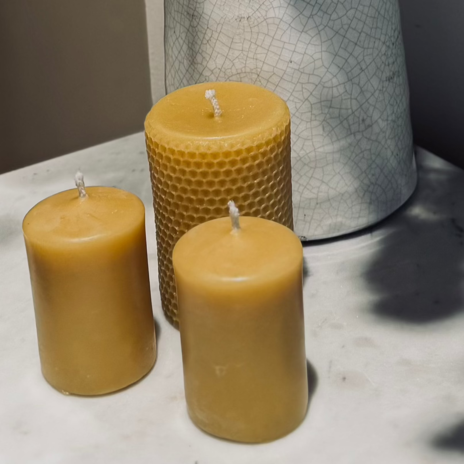 Two small natural, beeswax pillar candles, accompanied by a larger natural, beeswax pillar candle with a honeycomb imprint on the outside. They are positioned on a marble top table with a rustic milk jug behind and the shadow of some dried flowers surrounding it, with a neutral tone.