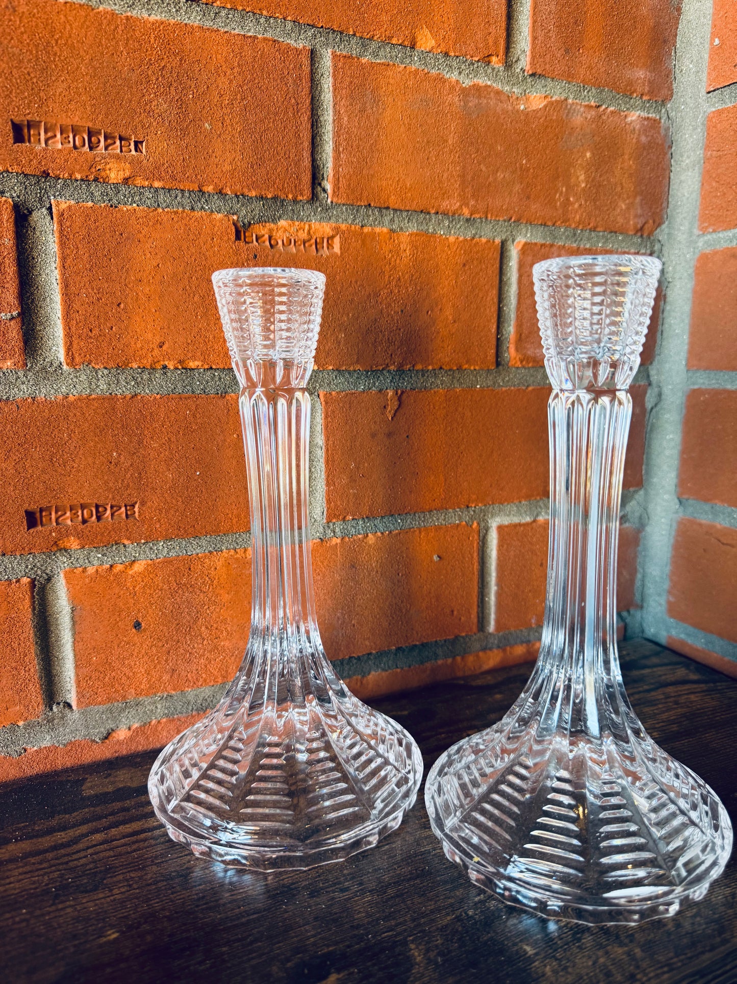 Two clear glass candle holders on a wooden surface with a brick wall background.