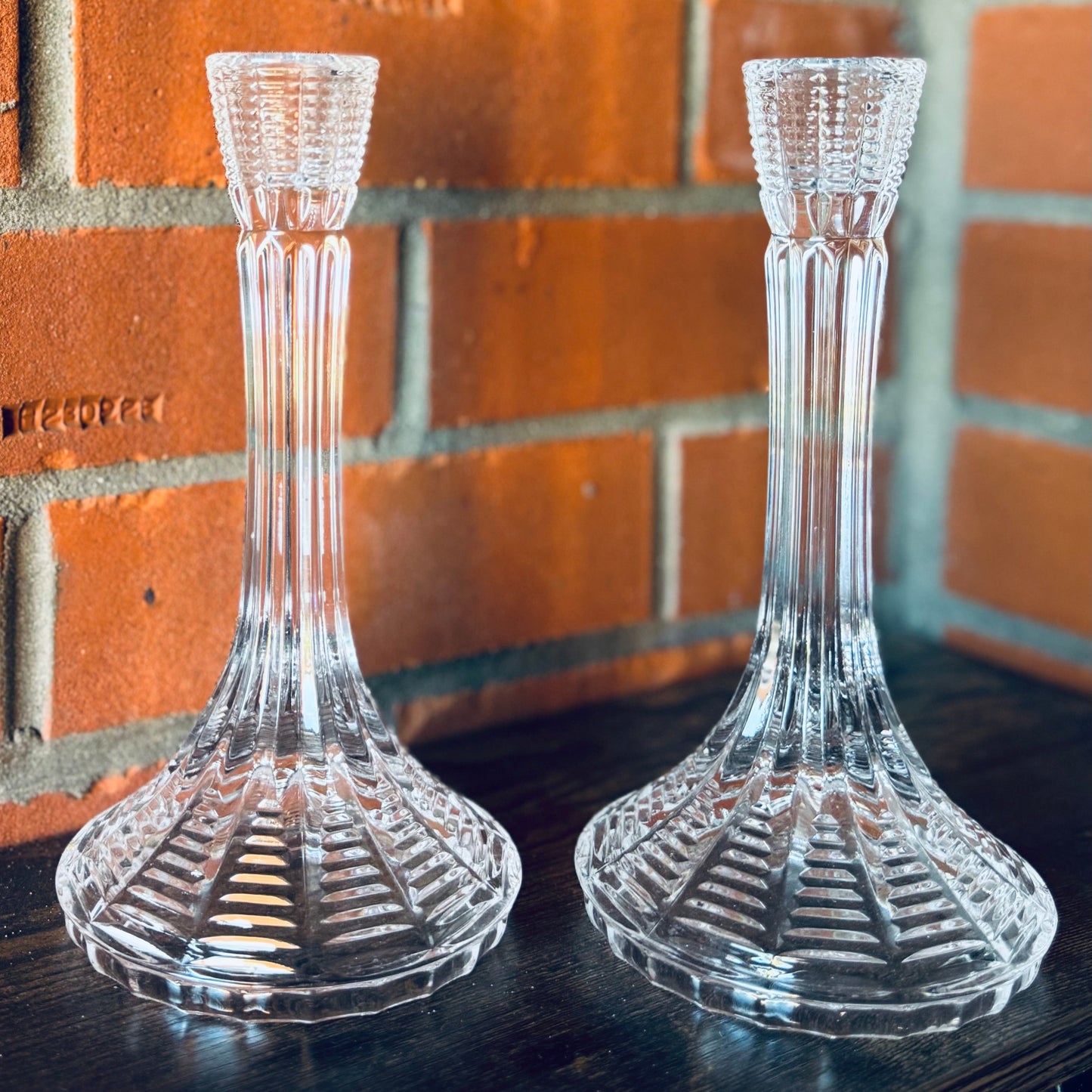 Two clear glass candle holders on a dark surface with a brick wall background