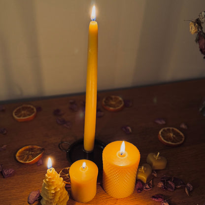 This shows a selection of lit beeswax candles, each one bringing a different unique charm. Positioned towards the back of the frame is a tall taper beeswax candle, with a charming christmas tree shaped candle to its left, and two different sized beeswax pillar candles to the right. There is a pair of unlit heart shaped tealight beeswax candles to the right, and at the front of the image, a lit, warming natural beeswax candle