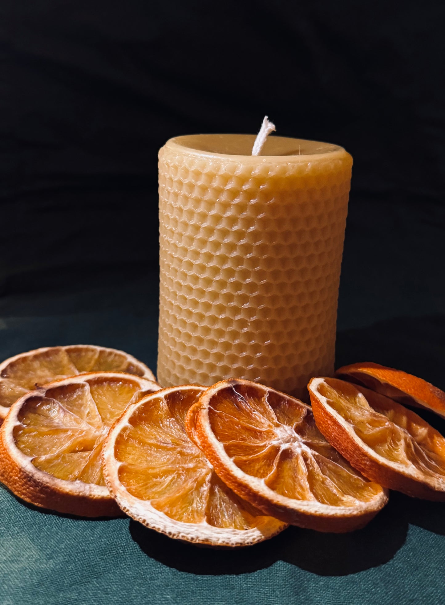 This image features a honeycomb imprinted beeswax candle which stands alone against a dark backdrop with dehydrated oranges surrounding the base. It shows a studio style lighting, allowing you to appreciate the honeycomb printing on the beeswax candle