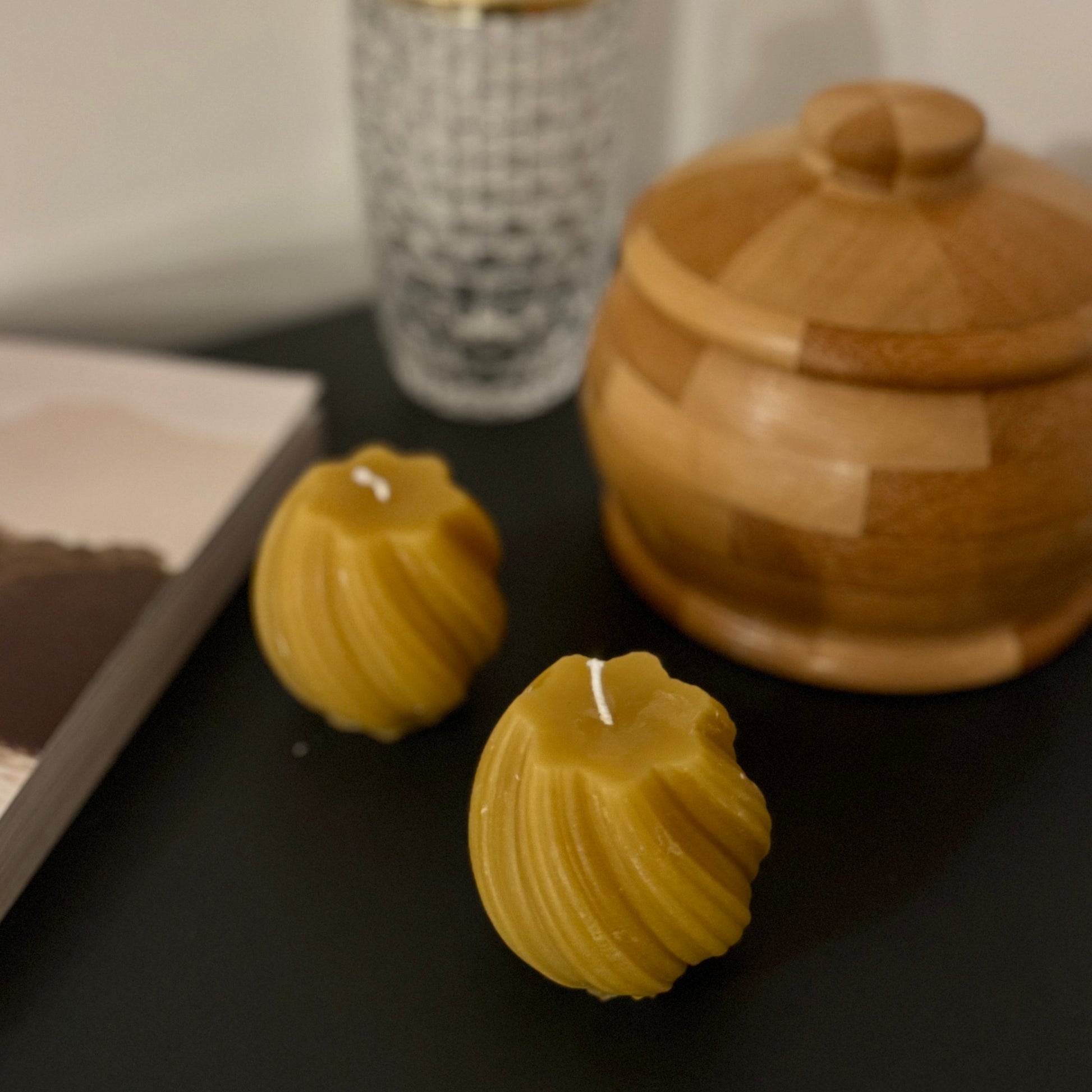 2 beeswax swirled candles sit on a grey sideboard, with some atural wooden products in the background, and a glass. The photograph is familiar and warm