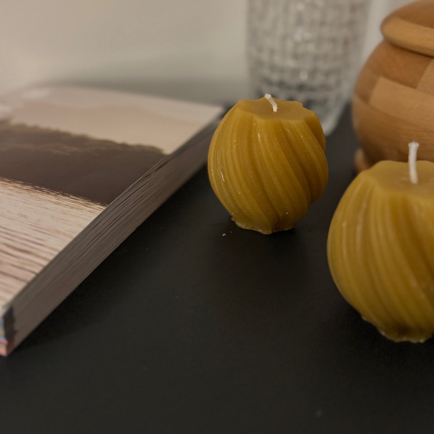 2 beeswax swirled candles sit on a grey sideboard, with some atural wooden products in the background, and a glass. The photograph is familiar and warm