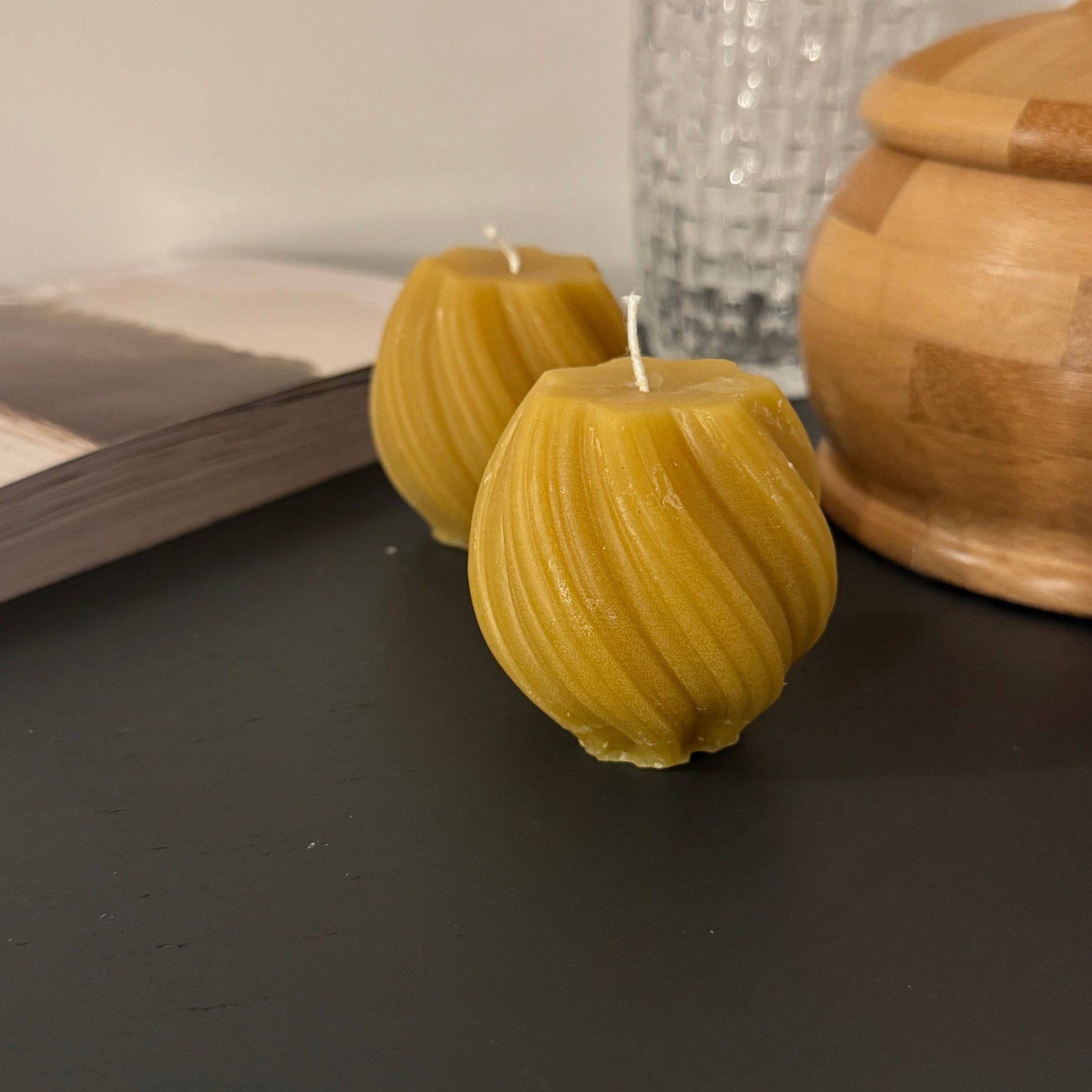 2 beeswax swirled candles sit on a grey sideboard, with some atural wooden products in the background, and a glass. The photograph is familiar and warm