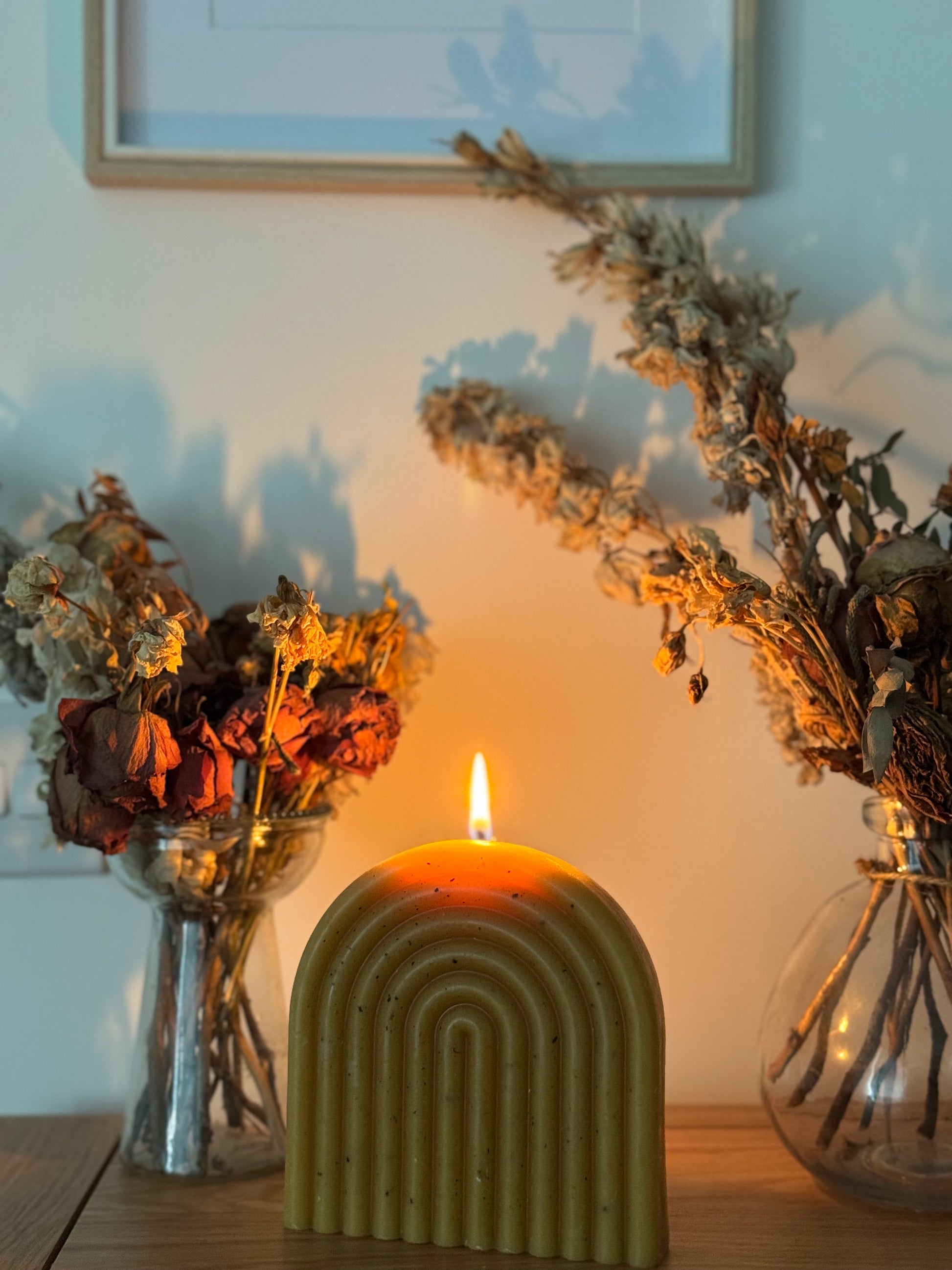 Decorative freckles beeswax candle with arch design on a table with dried flowers and a framed picture in the background.