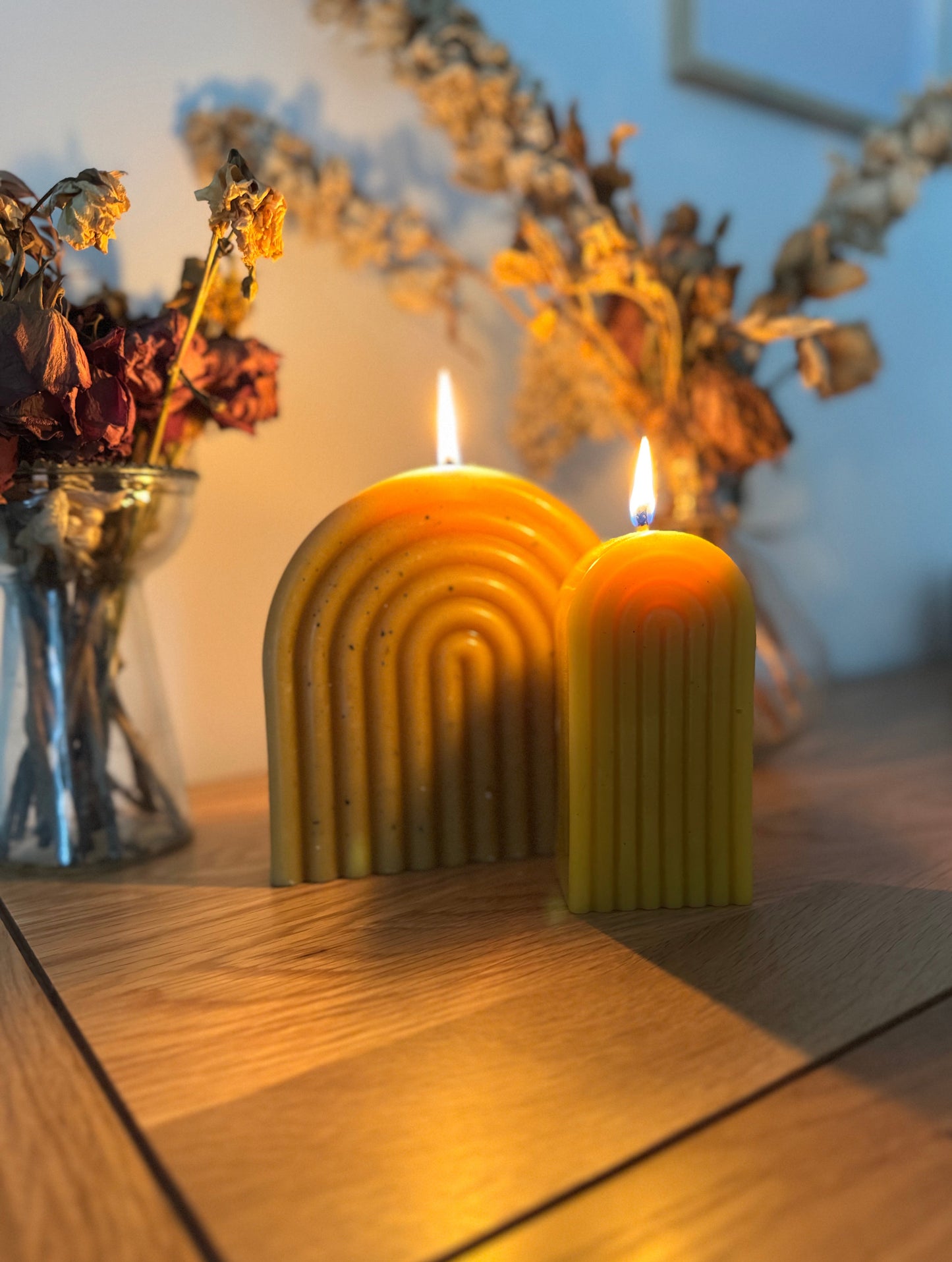 Two beeswax lit candles with a wavy design on a wooden surface, surrounded by dried flowers.