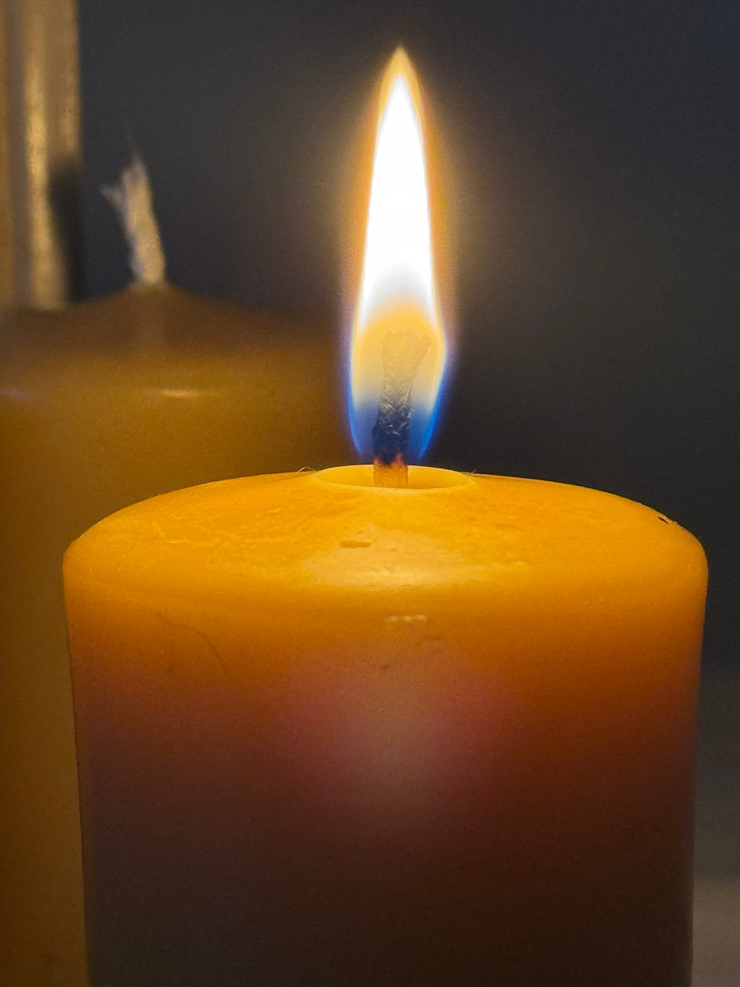 A natural beeswax candle stands lit in the centre of the frame, creating a cosy, soft ambience, with an unlit version behind. It is a close up of the flame and of the natural glow of beeswax candles. 