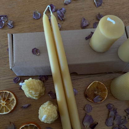 The image features a selection of natural beeswax candles, laid out on a rustic wooden table , adorned with dried rose petals and dehydrated oranges. It is a warm and inviting scene emphasising the natural allure of beeswax candles
