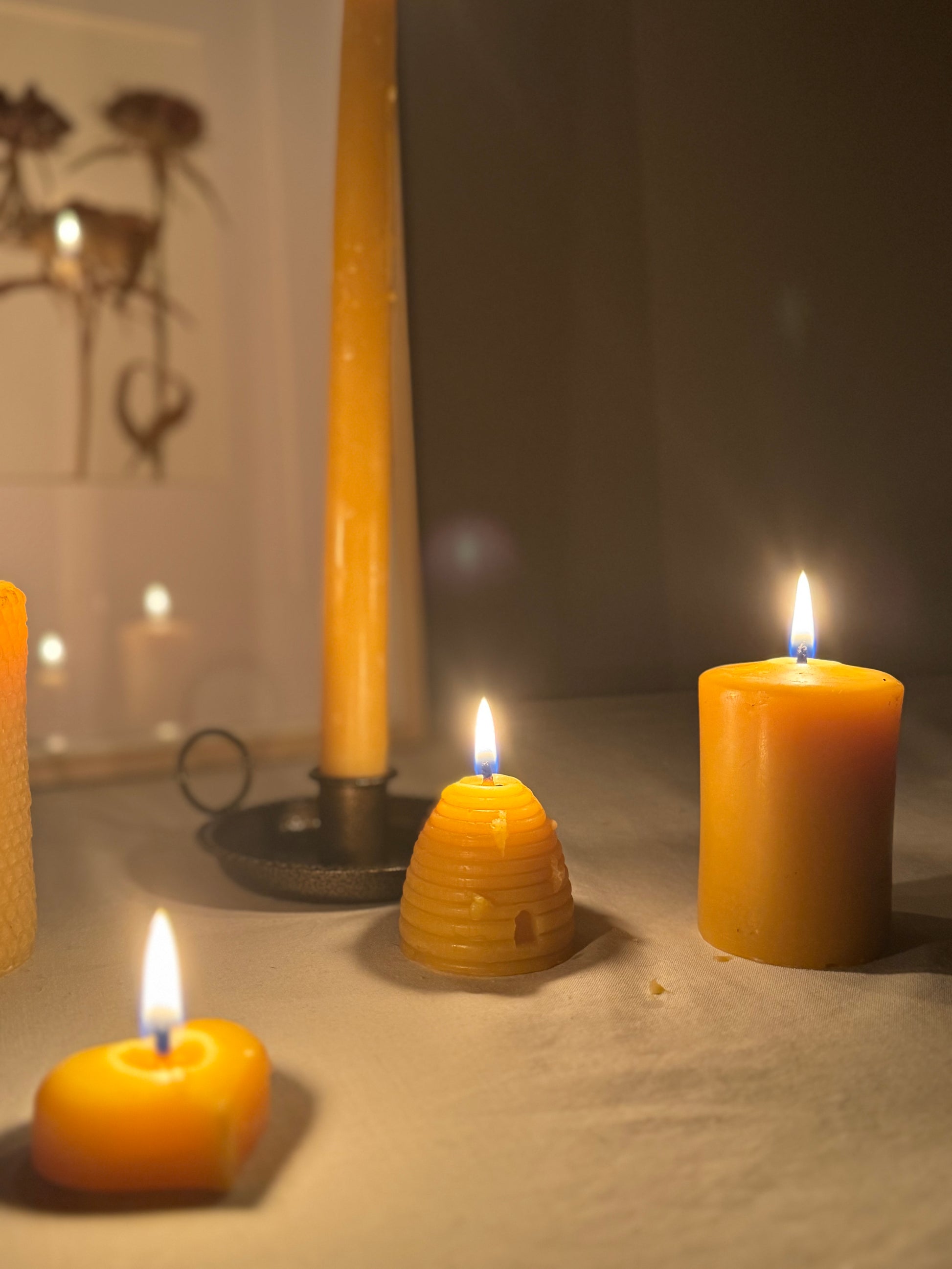 Four handcrafted beeswax candles displayed on a soft fabric surface, showcasing their natural golden hue and charming design. Perfect eco-friendly home decor with a warm, natural honey aroma and sustainable appeal. We see the base of a taper candle with the reflection shown in a dried flower frame situated behind. A small pillar candle, alongside a beehive-skep shaped candle, and a heart shaped beeswax tealight. 