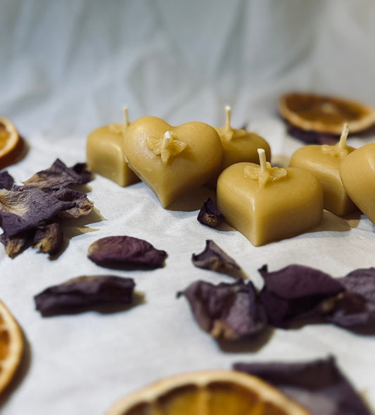 This image features a selection of natural beeswax heart shaped tealights displaed on a white backdrop with dried petals surround it for a natural, rustic and environmentally friendly appeal