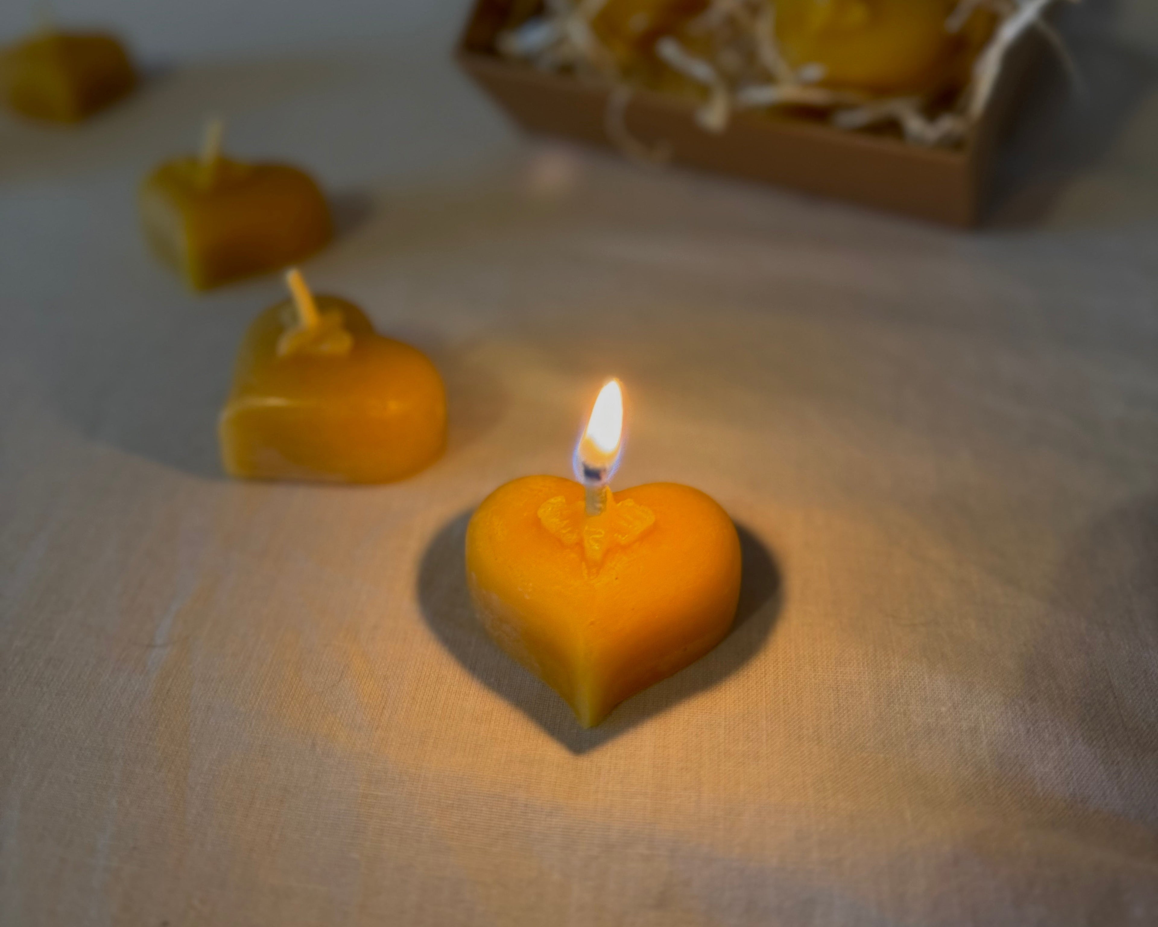 A glowing heart-shaped beeswax candle burning softly on a cream-colored tablecloth, surrounded by additional heart candles and a kraft box filled with natural shredded paper, creating a warm and cozy cosy ambiance