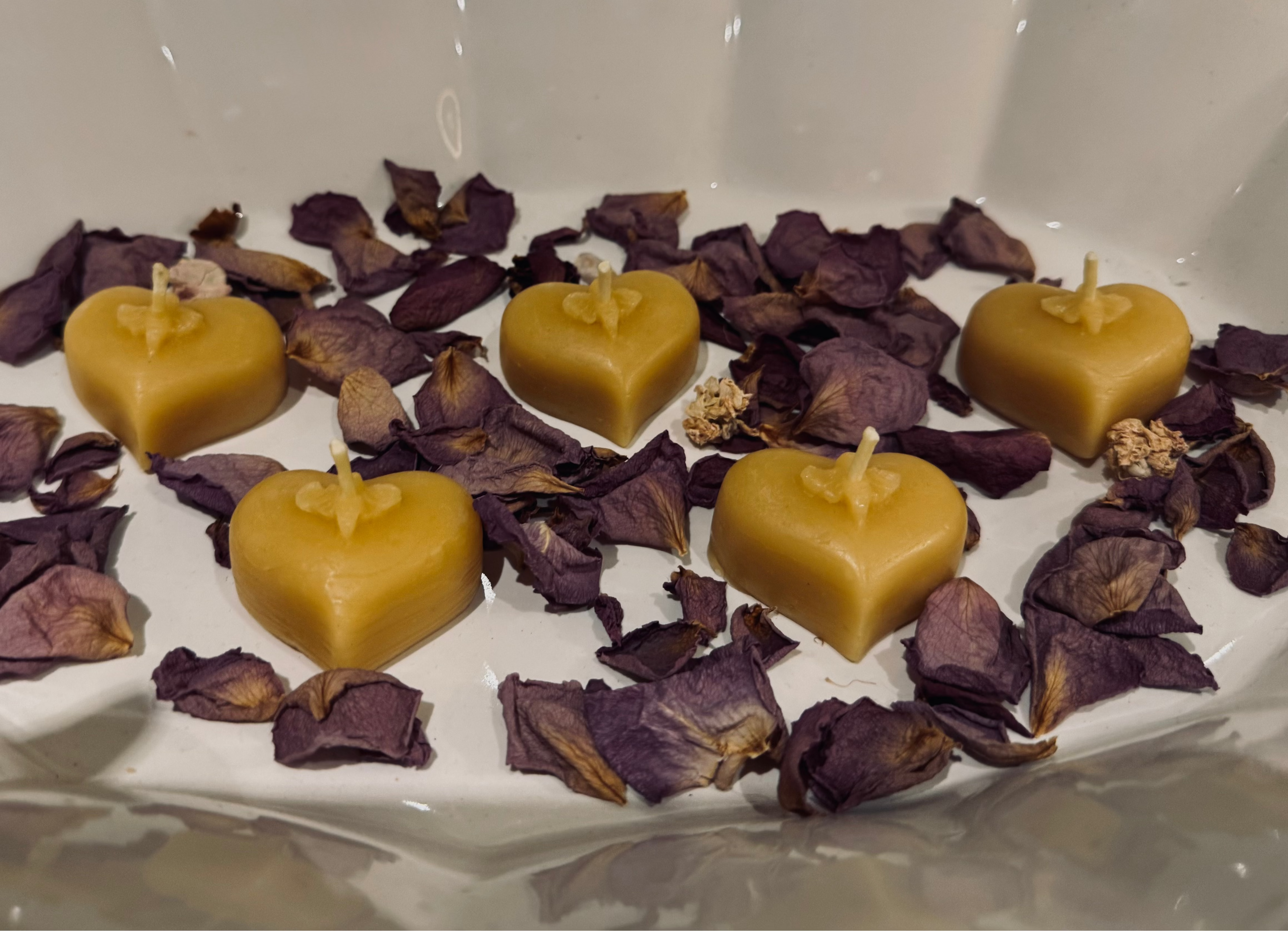 This image features 4 natural, beeswax heart tealights in a white ceramic bowl, accompanied by dried petals. The tealights have a cute bee motif on the top