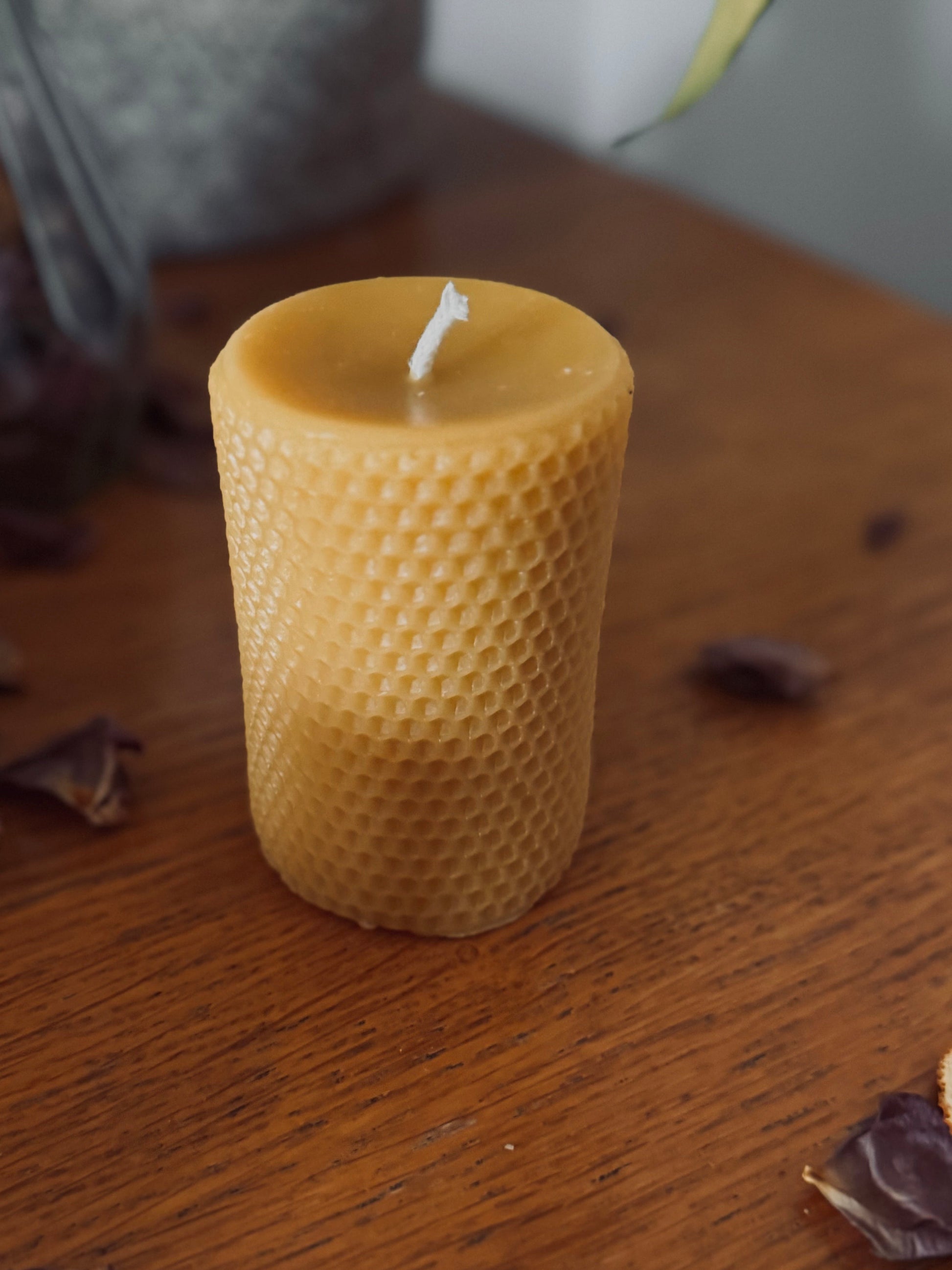 This image shows a natural beeswax candle with honeycomb printing on the outside. It's presented on a brown wooden backdrop with rustic aesthetic of dried petals and a hessian plant pot in the background. The ambiance is cosy and rustic, with a clear image of the beeswax natural candle