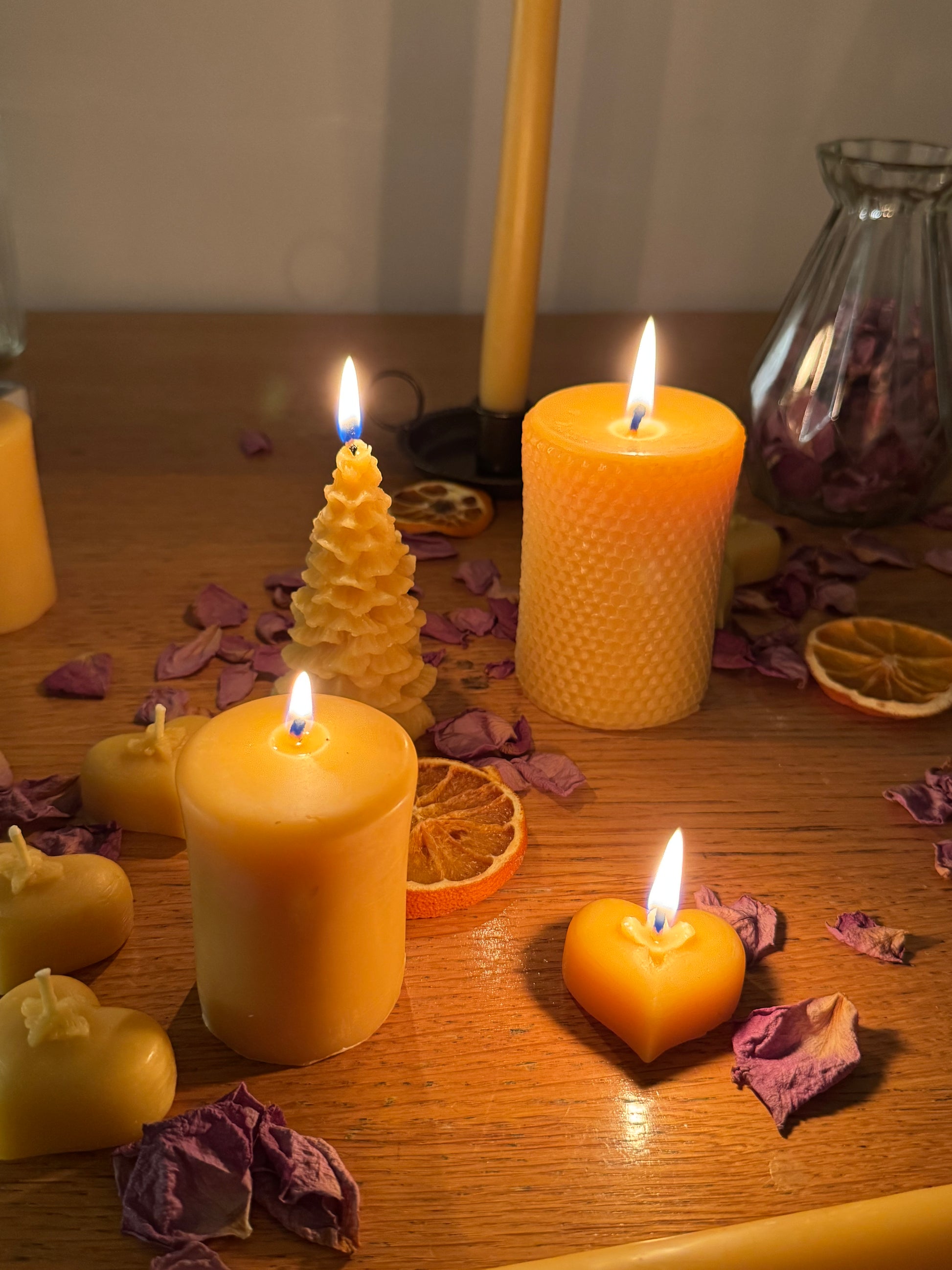 This image depicts beautiful lit natural beeswax candles in a range of forms, with a heart shaped natural beeswax tealight at the forefront, surrounded by dried petals and dehydrated oranges, creating a calm and tranquil, cosy vibe. The heart tealight is surroundedby a small pillar natural beeswax candle, a christmas tree shaped natural beeswax candle, and a honeycomb imprinted natural beeswax pillar candle, with the base of a taper beeswax candle in an iron candlestick holder behind.