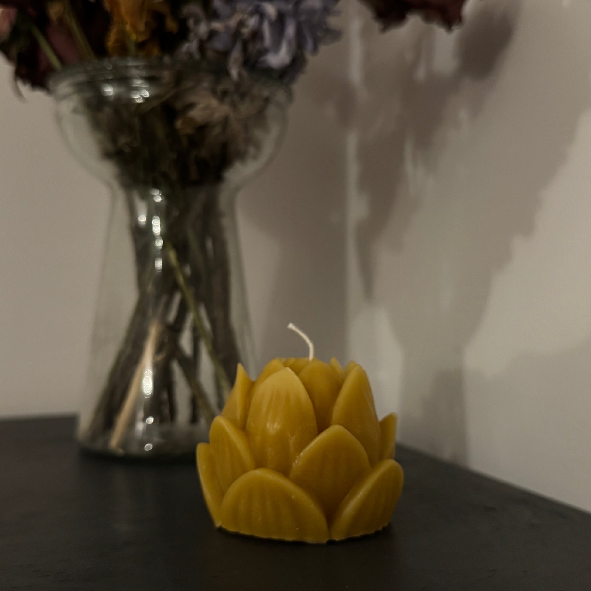 A singular beeswax candle,  positioned in the centre of the screen, with a vase of dried flowers behind, creating a warm, cosy, natural vibe