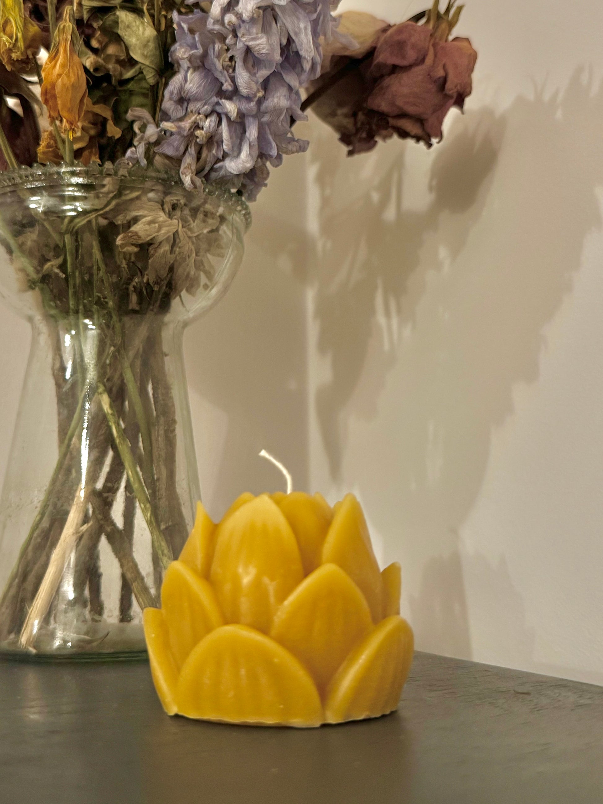 A singular beeswax candle,  positioned in the centre of the screen, with a vase of dried flowers behind, creating a warm, cosy, natural vibe