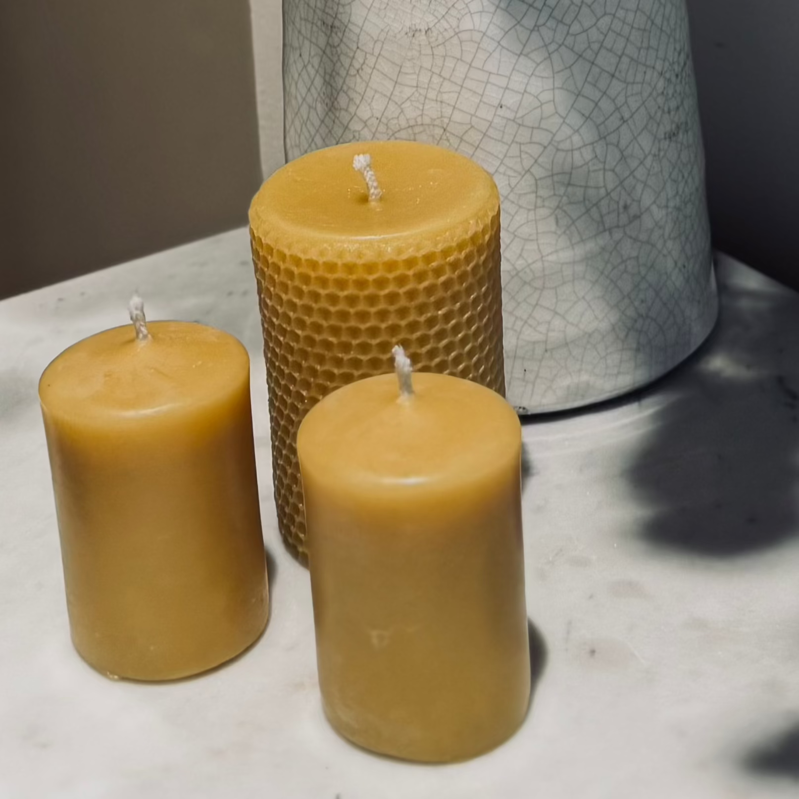 Two small natural, beeswax pillar candles, accompanied by a larger natural, beeswax pillar candle with a honeycomb imprint on the outside. They are positioned on a marble top table with a rustic milk jug behind and the shadow of some dried flowers surrounding it, with a neutral tone. 