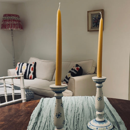 This image features two natural beeswax taper candles in a home setting with blue candlesticks and a blue runner across a rustic country table