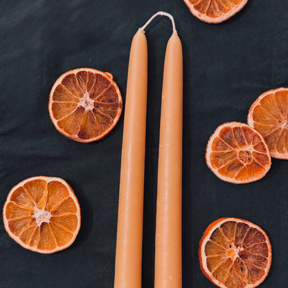 The image features a natural beeswax taper candle laying on a dark green backdrop with dehydrated oranges around. The wicks are connecting, creating a rustic vibe and truly handmade feel
