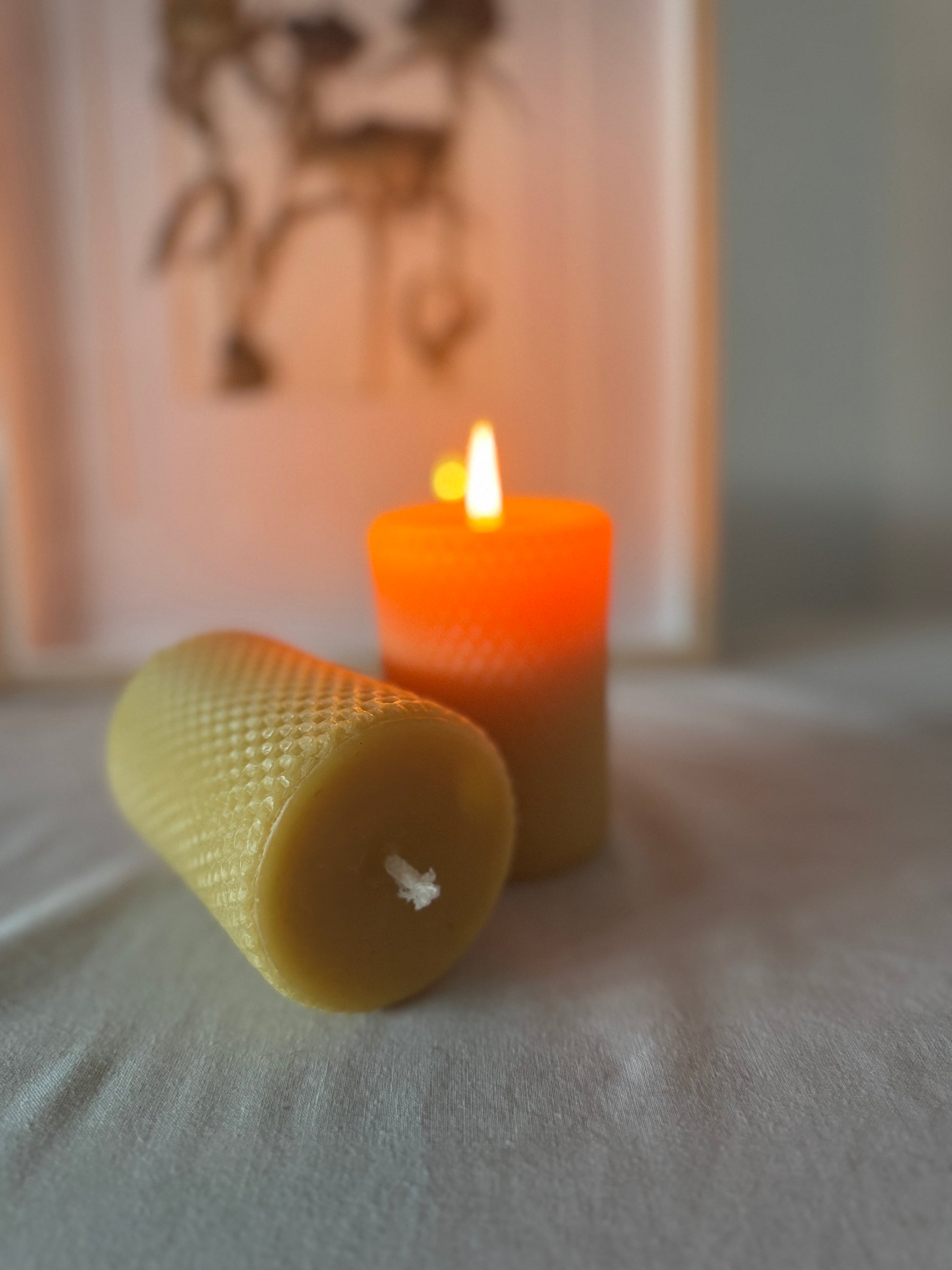 This image shows a natural beeswax candle with honeycomb printing on the outside which is unlit, laying in front of a lit beeswax hexagonal candle which is lighting the background. It's presented on a white linen backdrop with rustic aesthetic of dried flower in the background. The ambiance is cosy and rustic, with a clear image of the beeswax natural candles whilst being used and not in use.