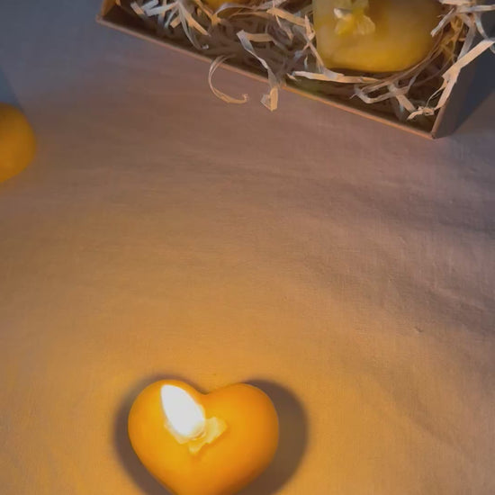 A lit cosy, natural beeswax candle lists in the forefront of the video, flickering gently, with a  pair of heart shaped tealights sit behind packaged in eco-friendly kraft packaging with a recyclable wood wool base