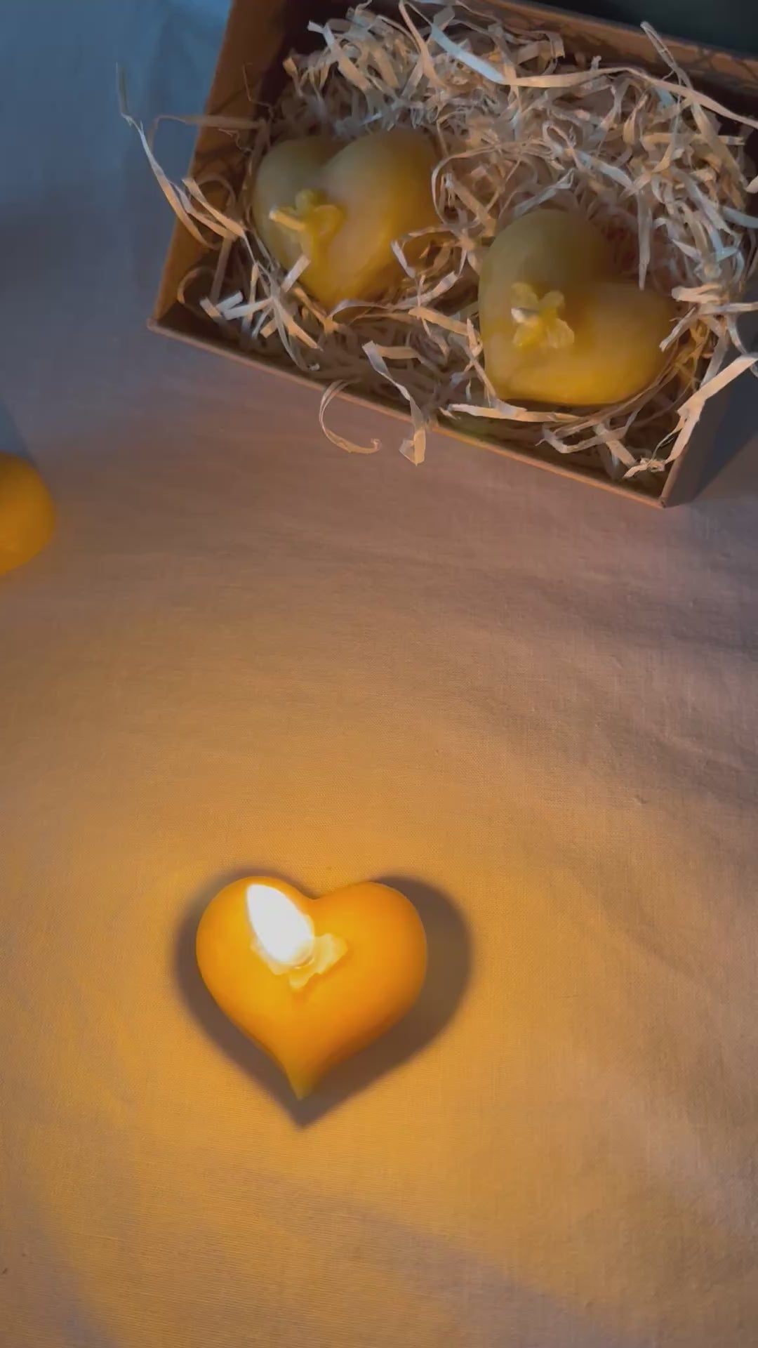 A lit cosy, natural beeswax candle lists in the forefront of the video, flickering gently, with a  pair of heart shaped tealights sit behind packaged in eco-friendly kraft packaging with a recyclable wood wool base